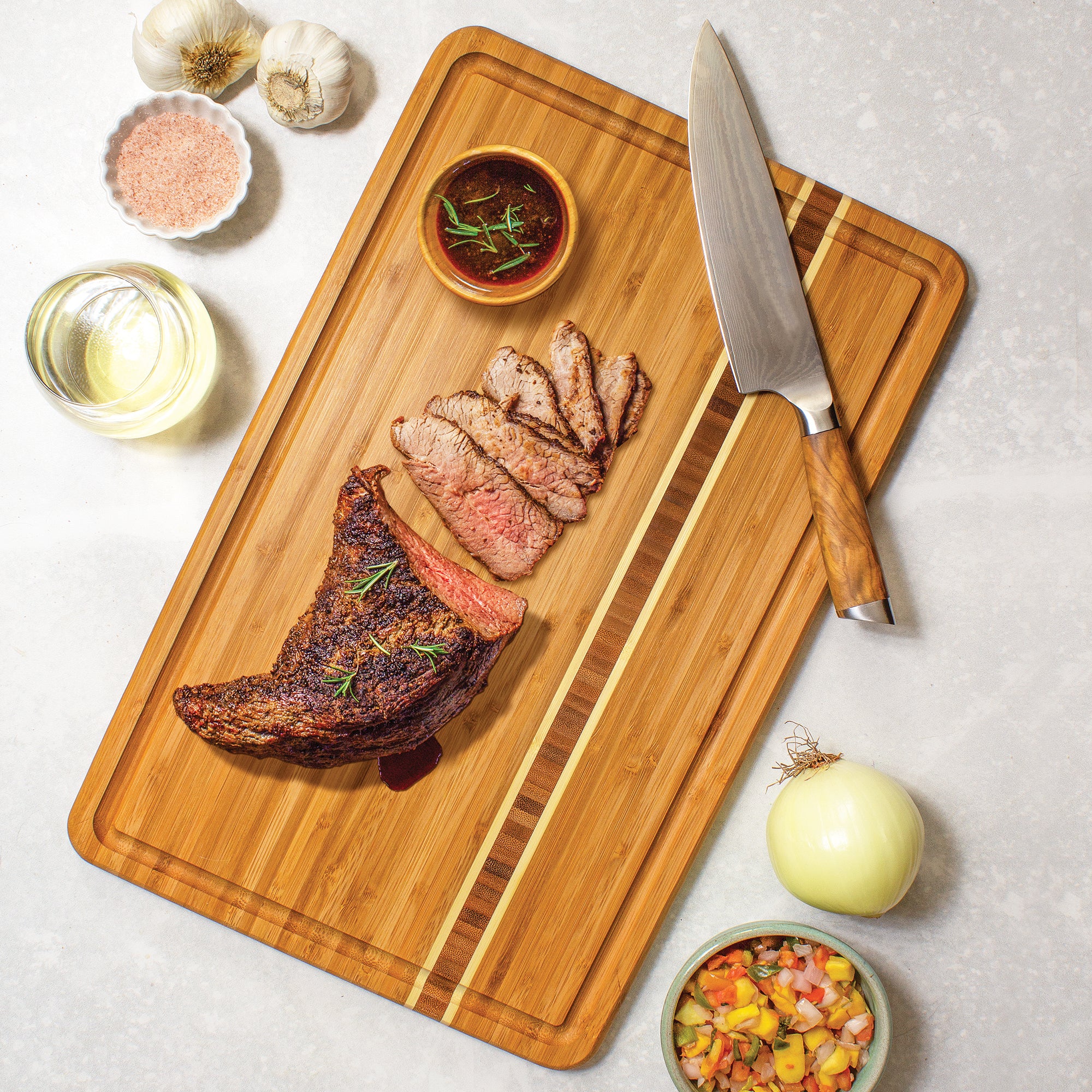 The Dominica Carving Board by Totally Bamboo, from the Caribbean Collection, showcases sliced, cooked steak with rosemary. It’s surrounded by a sauce bowl, knife, onions, mango salsa, garlic, salt, and wine on a marble surface. Dimensions: 20-5/8" x 12-1/2".