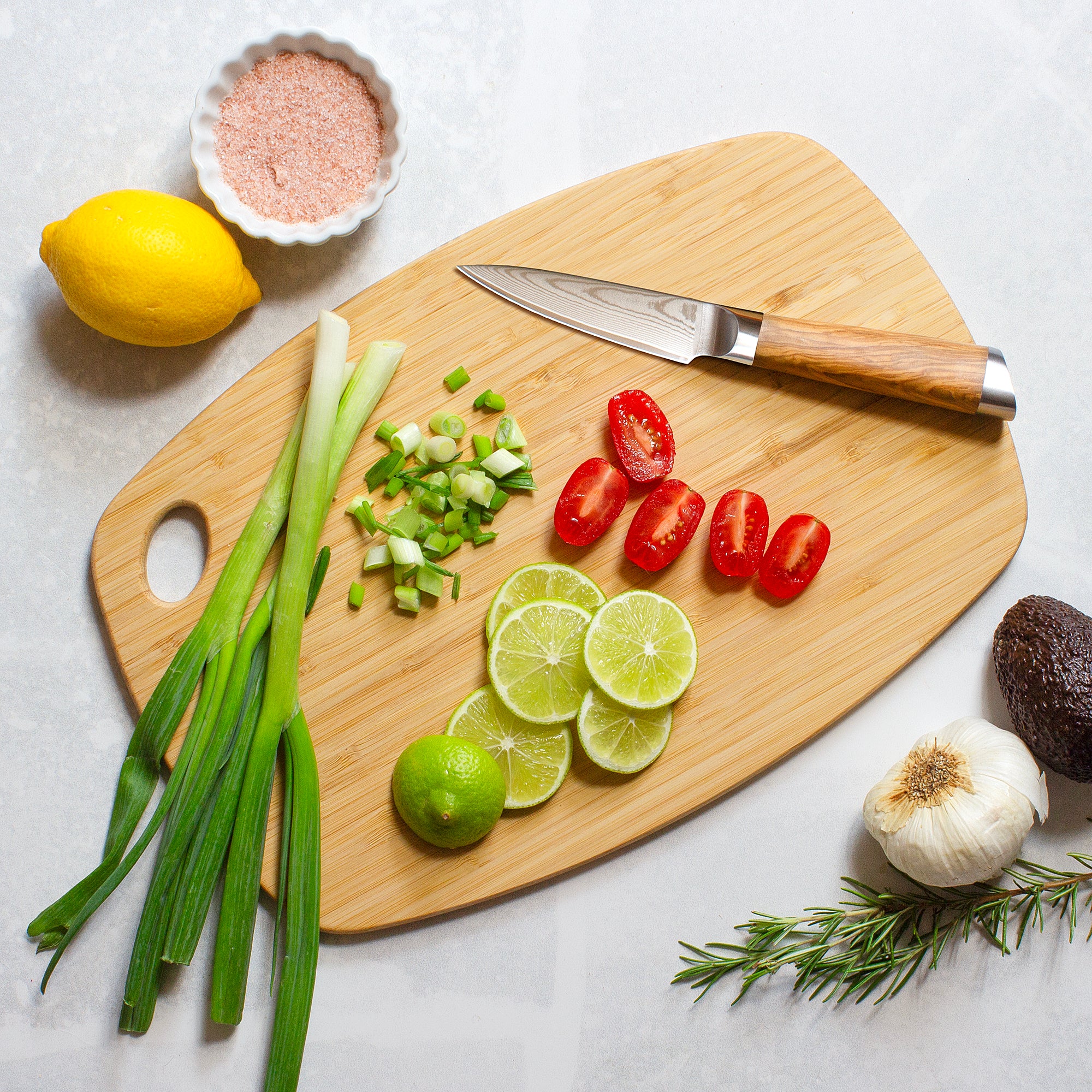 The Totally Bamboo GreenLite™ Dishwasher Safe Cutting Board, 13-1/2" x 9", holds a knife, sliced green onions, grape tomatoes, and lime slices for the perfect kitchen prep scene on a light surface.